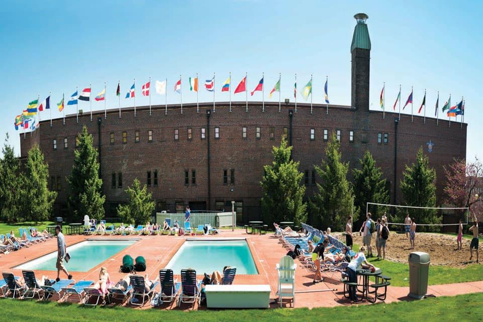 People play sand volleyball and lie on chairs next to two swimming pools.