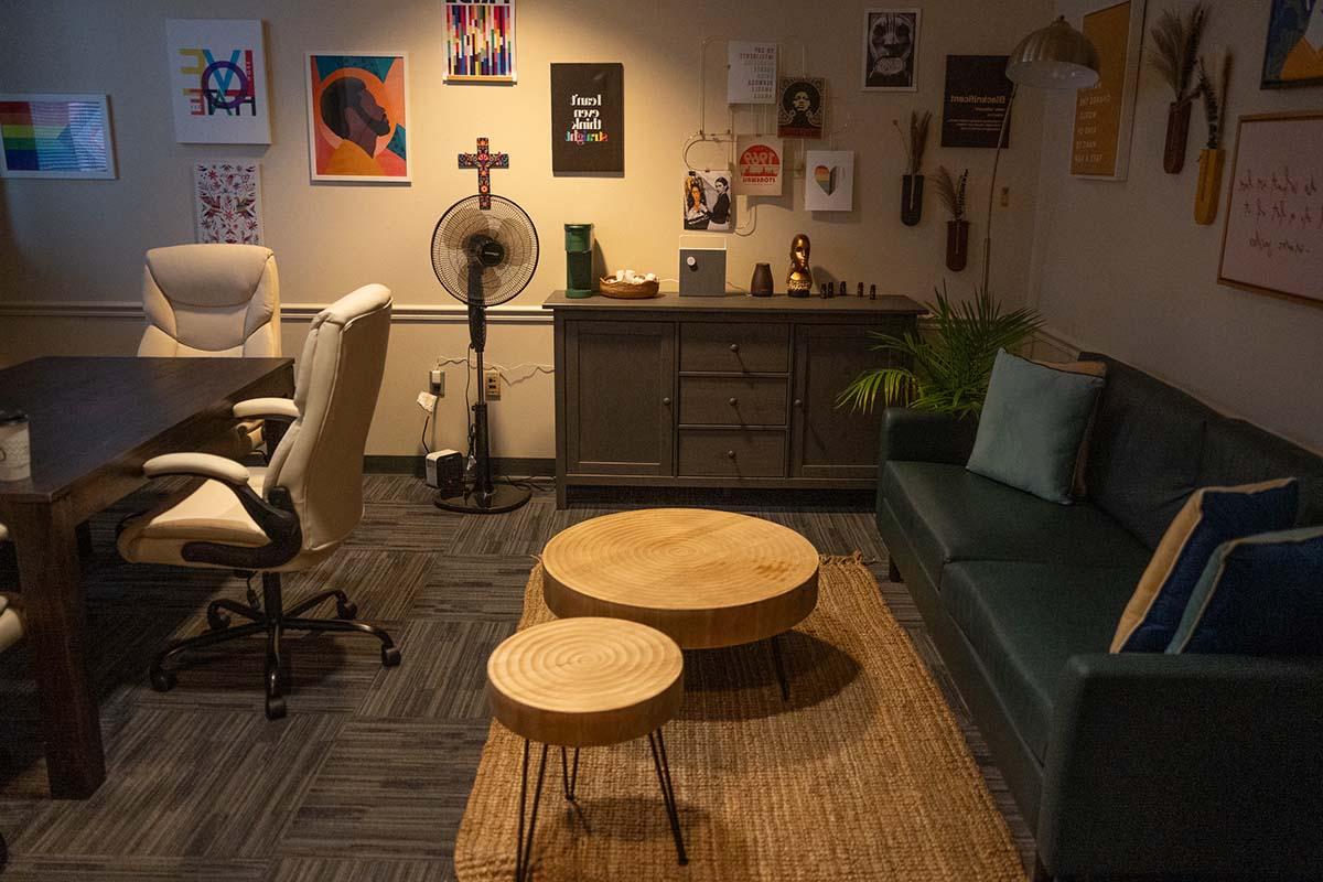 Image of a room with low lighting, a table and chairs, sofa, woven floor rug and round coffee tables, with posters on the wall.