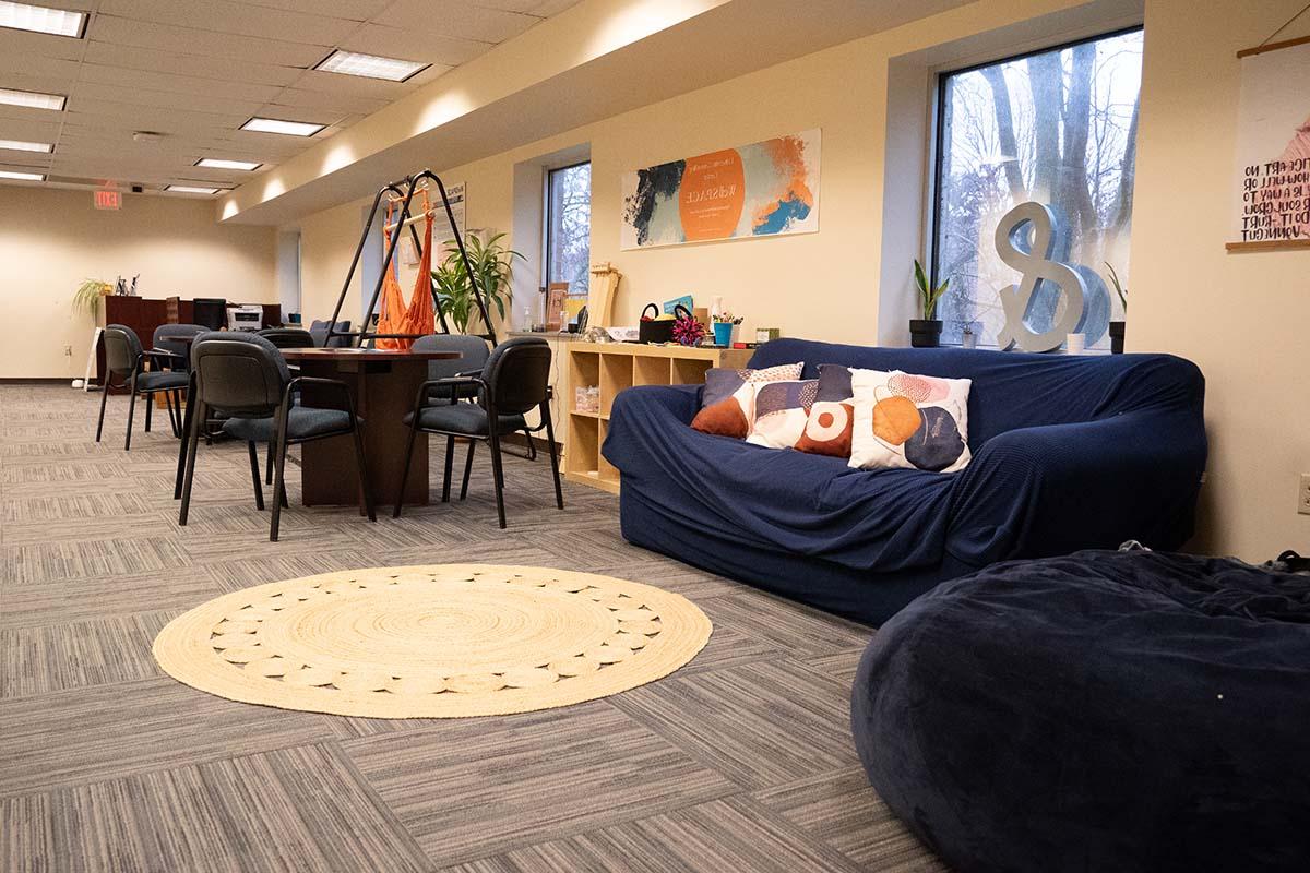 Image of a room with carpeting, a round area rug, a comfy looking couch, bean bag chair, and tables with chairs.